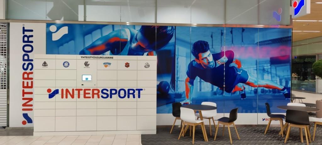 Intersport Karisman kaupan julkisivu, jossa kuvat urheilevista naisesta ja miehestä ja niiden edessä vaalea Intersport logolla varustettu pakettiautomaatti.